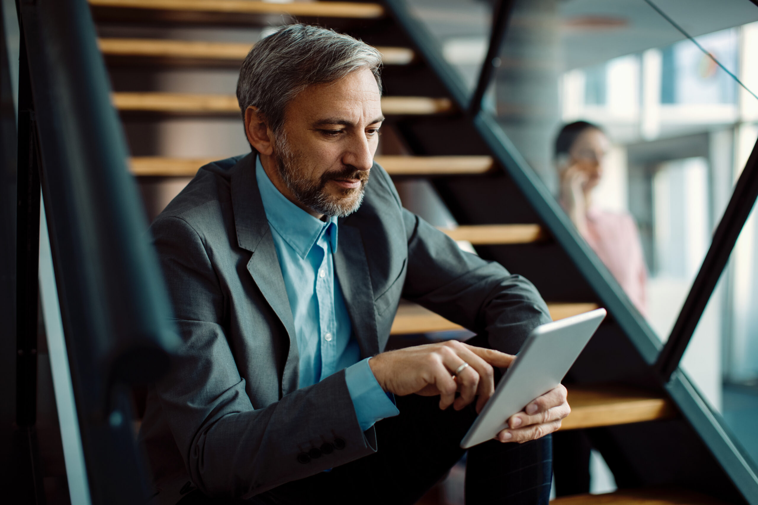 Mid adult businessman surfing the net on touchpad while relaxing on staircase in the office.
